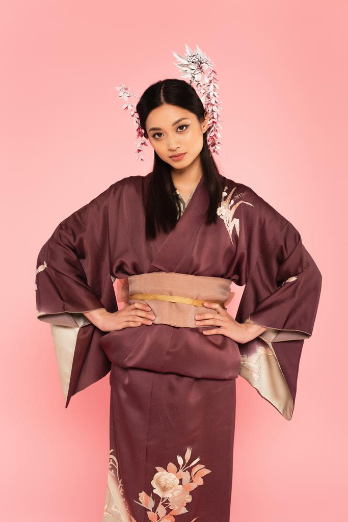 Joven mujer asiática en ropa tradicional cogida de la mano en las caderas aislado en rosa  - Foto, Imagen