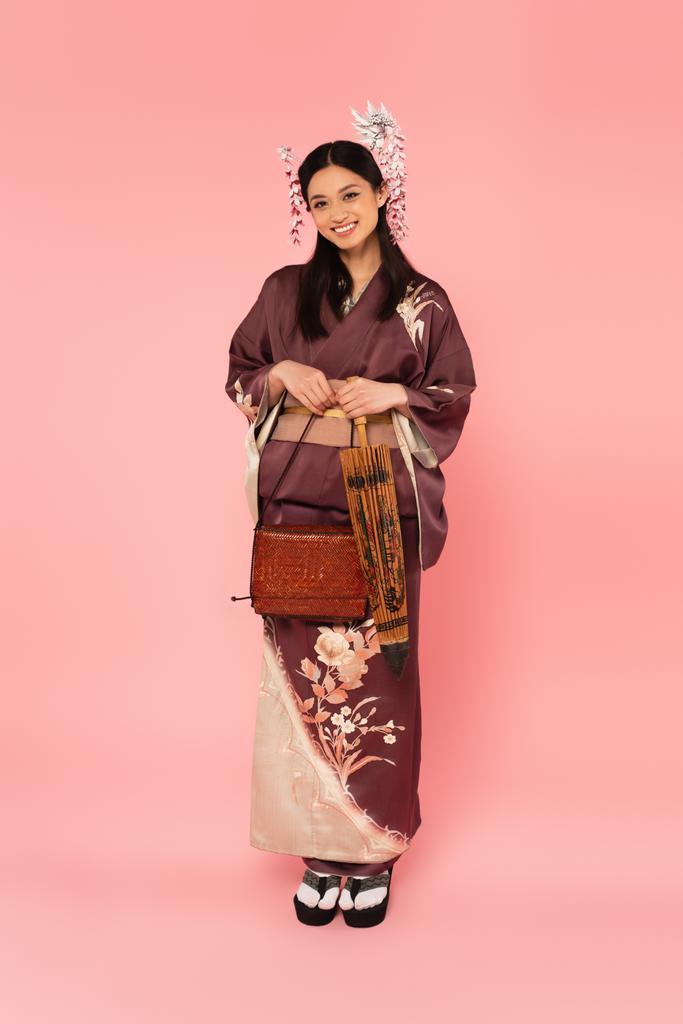 Mulher japonesa em quimono segurando guarda-chuva e bolsa no fundo rosa - Foto, Imagem