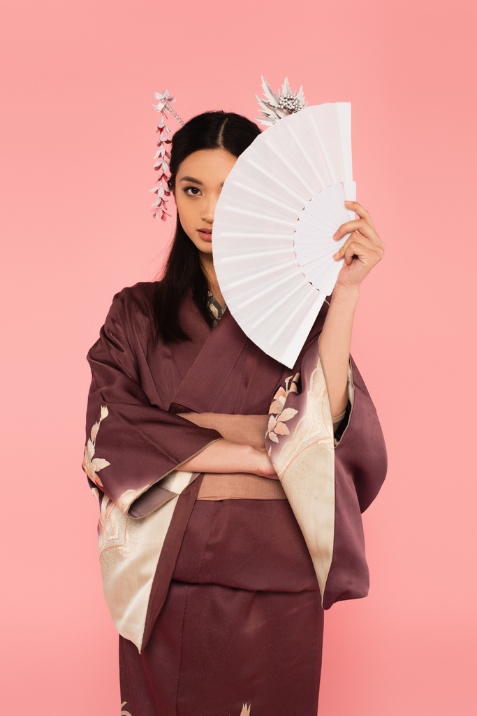 Азиатка в кимоно держит вентилятор рядом с лицом изолирован на розовый  - Фото, изображение