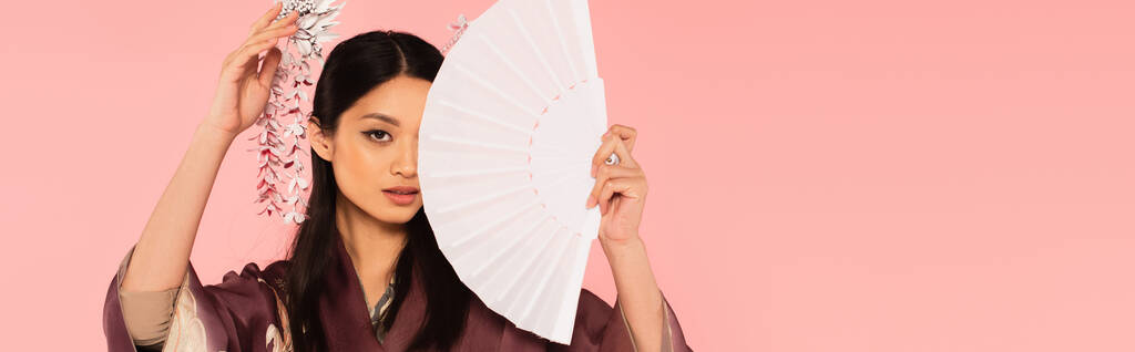 Asian woman with traditional hairdo holding fan near face isolated on pink, banner  - Photo, Image