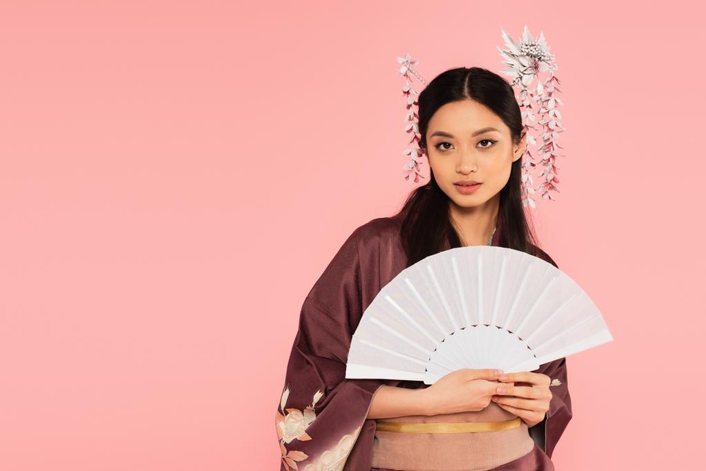 Csinos ázsiai nő kezében fehér ventilátor és nézi kamera elszigetelt rózsaszín  - Fotó, kép