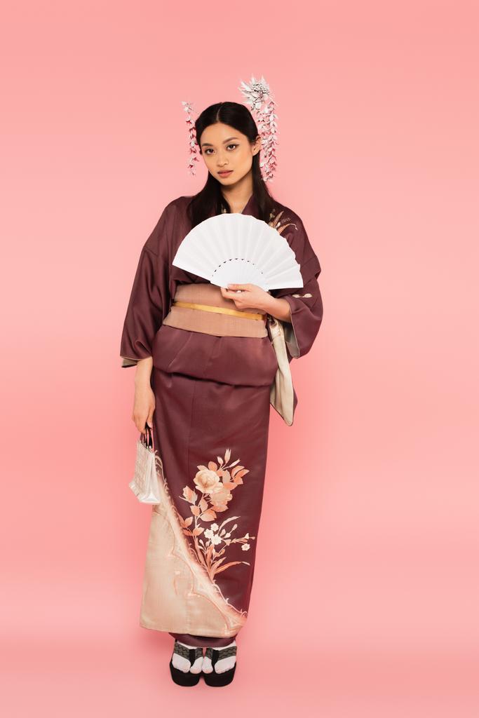 Pełna długość azjatyckiej kobiety z torebką i wentylatorem stojącej na różowym tle - Zdjęcie, obraz
