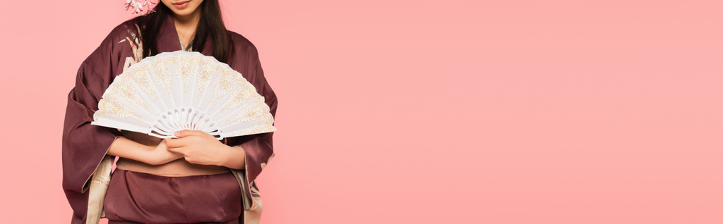 Обрізаний вид жінки в східному кімоно тримає вентилятор ізольовано на рожевому, банер
  - Фото, зображення