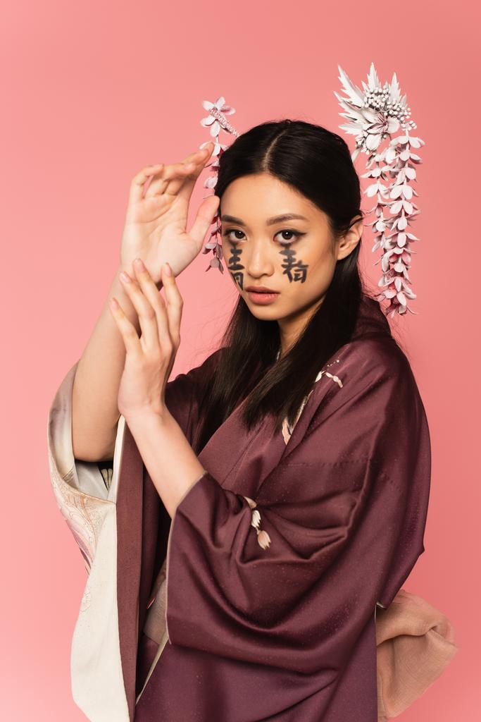 Γιαπωνέζα με ανατολίτικα ρούχα και ιερογλυφικά στο πρόσωπο, απομονωμένη σε ροζ.  - Φωτογραφία, εικόνα