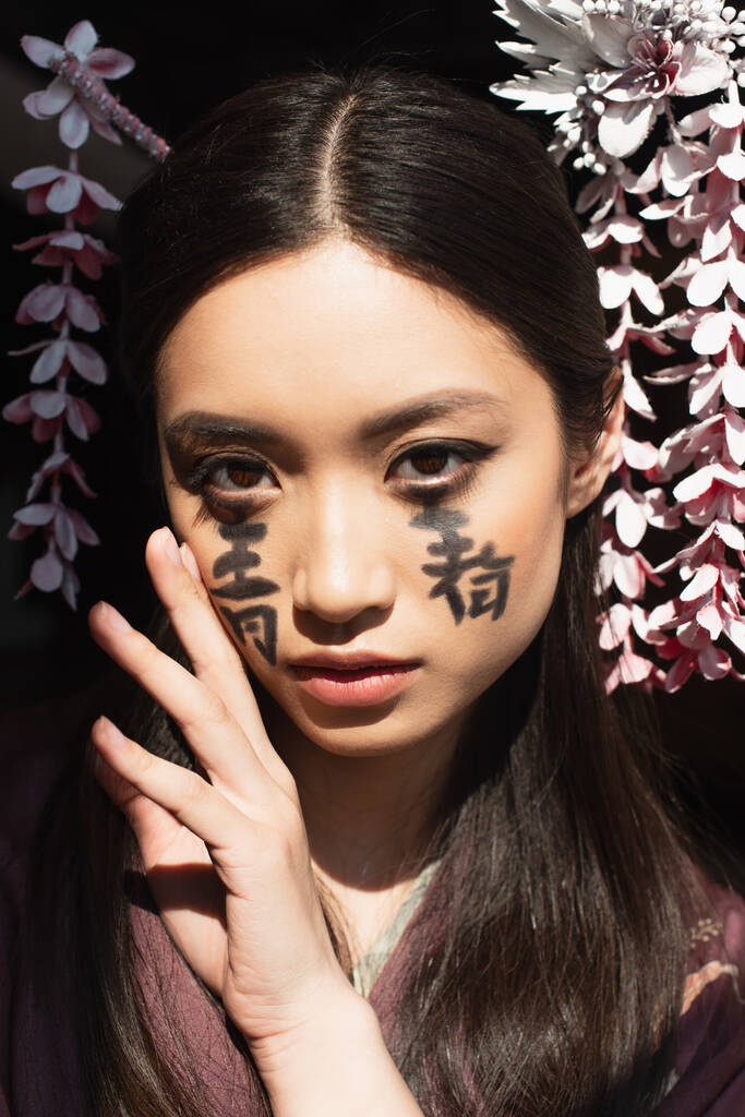Mooie aziatische vrouw met hiërogliefen op gezicht en haarsticks geïsoleerd op zwart  - Foto, afbeelding