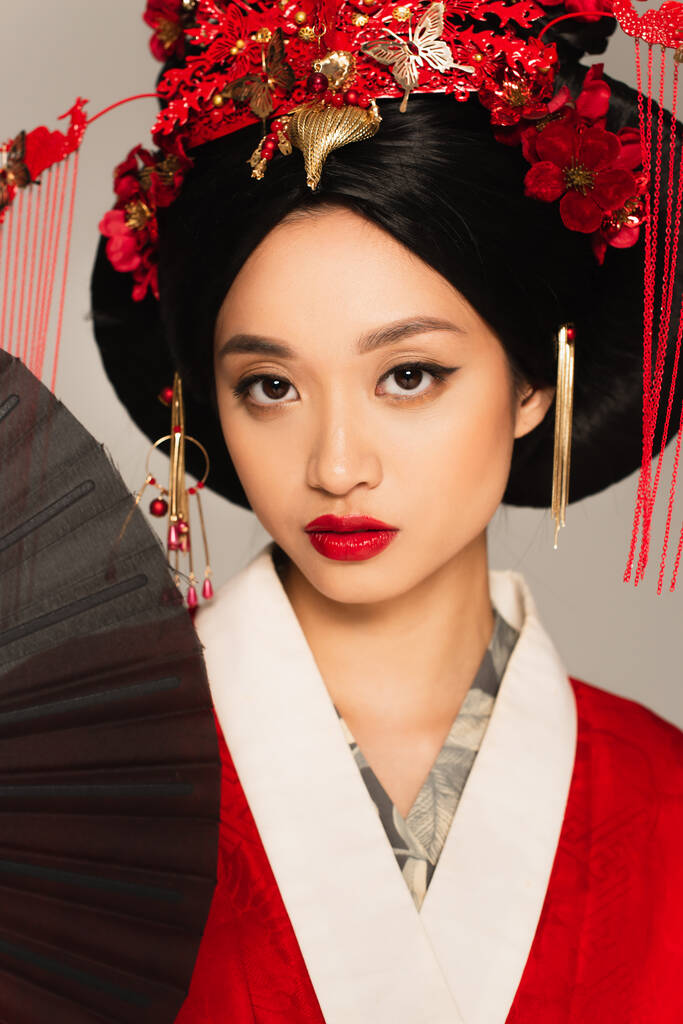 Femme asiatique avec lèvres rouges tenant ventilateur noir isolé sur gris  - Photo, image