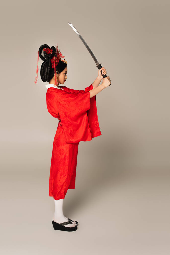 Zijaanzicht van japanse vrouw in rode kleren kijken naar zwaard op grijze achtergrond  - Foto, afbeelding
