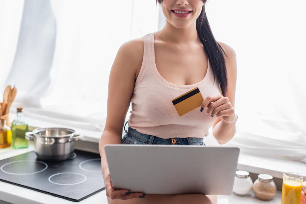 καλλιεργημένη άποψη της νεαρής γυναίκας που κατέχει πιστωτική κάρτα και τη χρήση φορητού υπολογιστή στην κουζίνα  - Φωτογραφία, εικόνα
