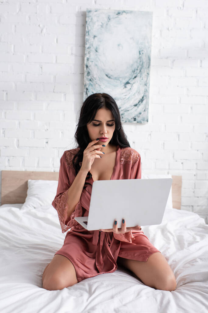 junge Frau im Seidenmantel hält Laptop im Schlafzimmer  - Foto, Bild