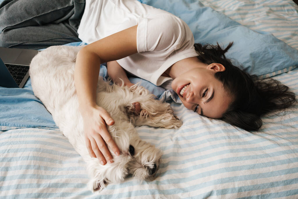 Boldog fehér nő simogatja a kutyáját, miközben otthon fekszik az ágyon. - Fotó, kép
