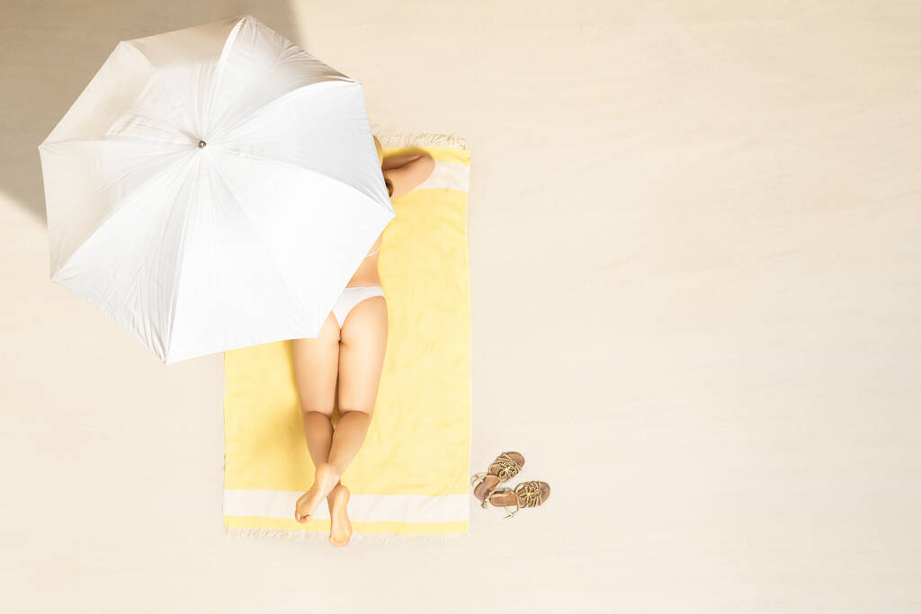 Sama žena v bikinách sedící a opalující se na žlutém plážovém ručníku. pod deštníkem. Žena relaxace na písečné pláži o letních prázdninách. Horní, letecké zobrazení - Fotografie, Obrázek