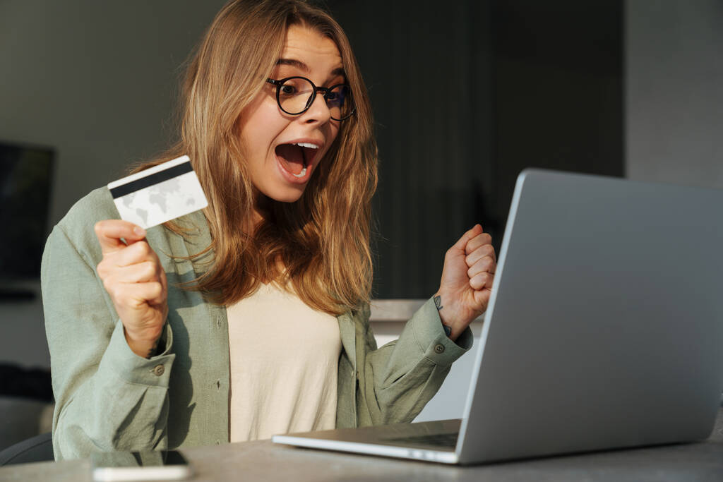 Belle femme excitée tenant la carte de crédit et travaillant avec un ordinateur portable à la maison - Photo, image