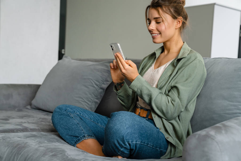 Счастливая милая женщина, пользующаяся мобильным телефоном, сидя дома на диване - Фото, изображение