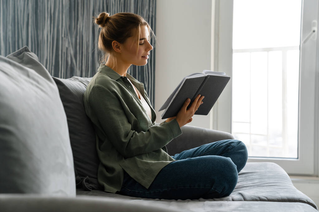 Onnellinen houkutteleva nuori nainen lukemassa kirjaa istuessaan sohvalla kotona - Valokuva, kuva