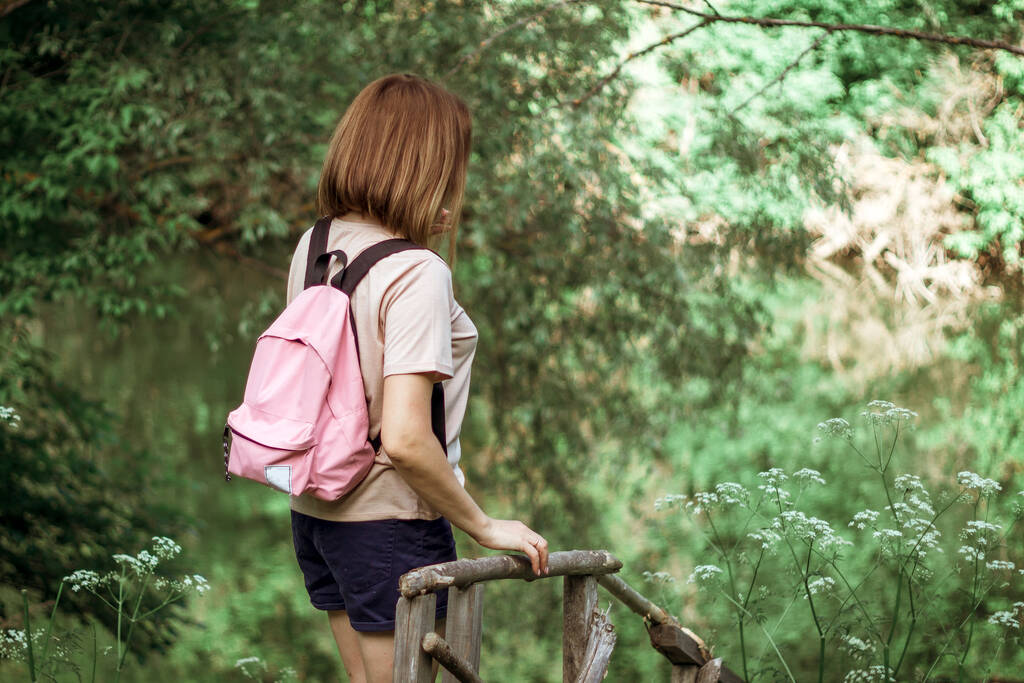 Nuori nainen, jolla on lyhyet hiukset ja vaaleanpunainen reppu, seisoo selkä kameralle pienellä puisella sillalla lähellä jokea metsässä. Valikoiva fokus. - Valokuva, kuva
