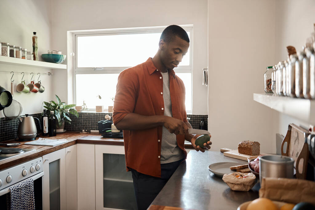 Junger Afrikaner halbiert Avocado, während er zu Hause an der Theke in seiner Küche steht - Foto, Bild
