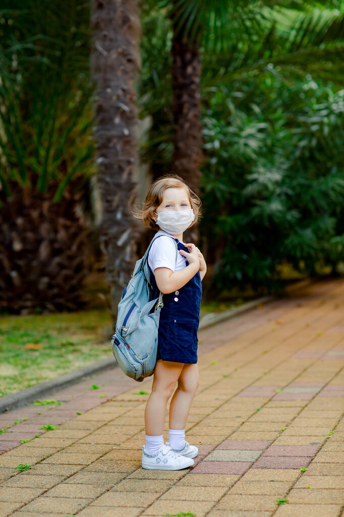 pieni tyttö, koulutyttö, menee kouluun puistossa denim-reppu ja lääketieteellinen naamio - Valokuva, kuva