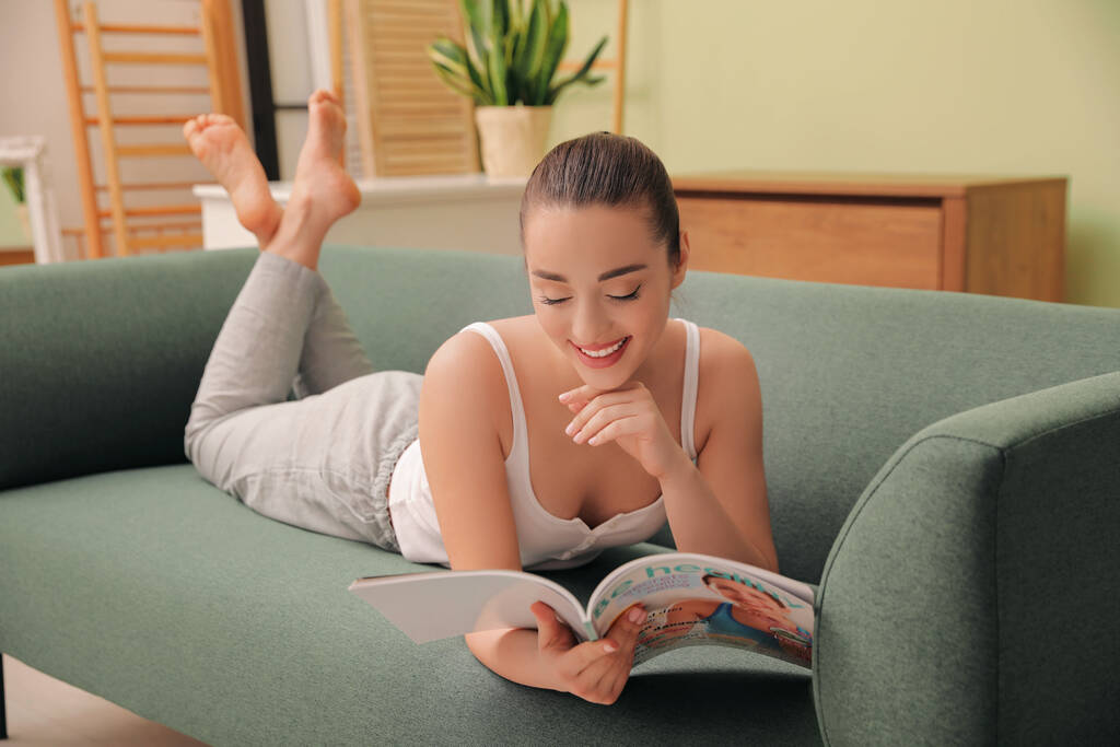 Happy woman reading magazine on sofa at home - Photo, Image