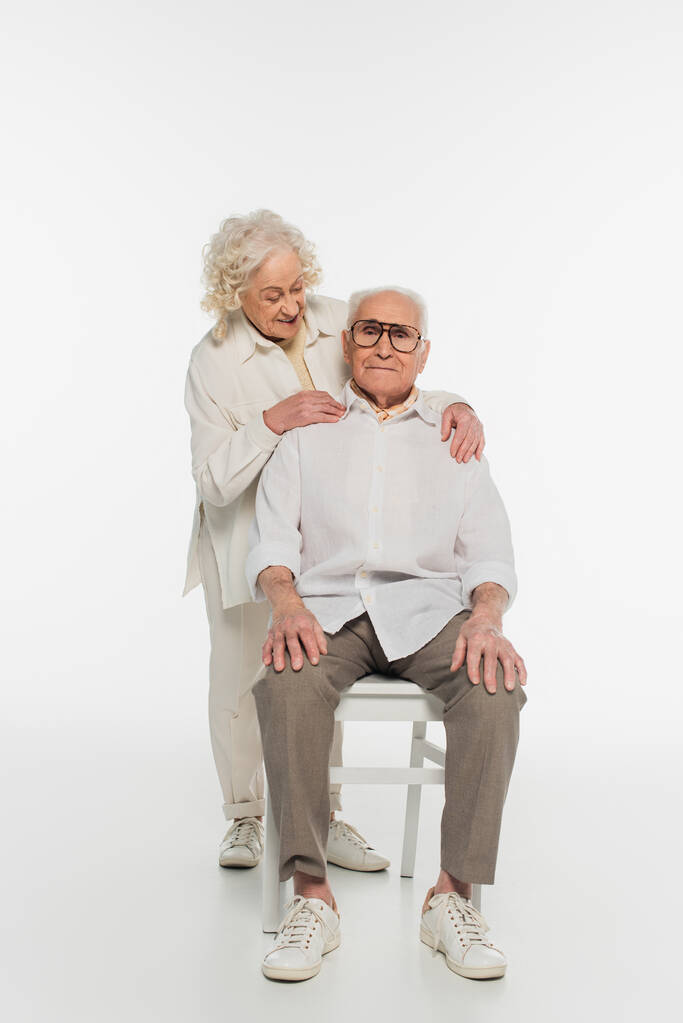 ηλικιωμένη γυναίκα αγκαλιάζει απαλά τον σύζυγο που κάθεται στην καρέκλα στα λευκά - Φωτογραφία, εικόνα