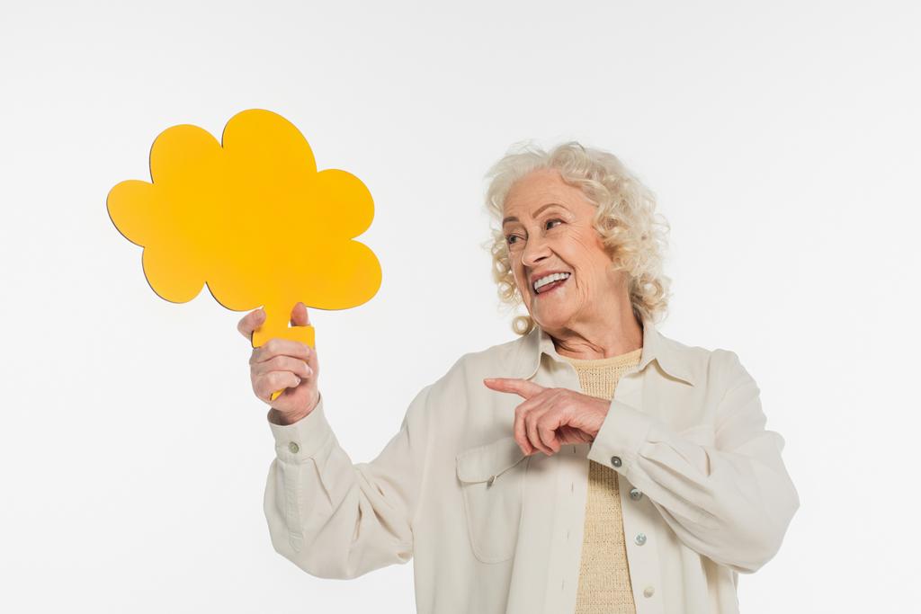 улыбающаяся пожилая женщина, указывающая пальцем на желтый мыслепузырь в руке, изолированный на белом - Фото, изображение