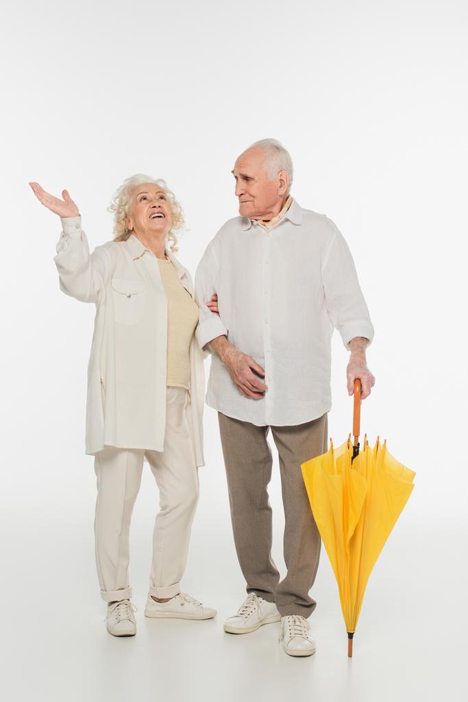 verbaasd oudere vrouw met de hand in de lucht staan met echtgenoot houden paraplu op wit - Foto, afbeelding