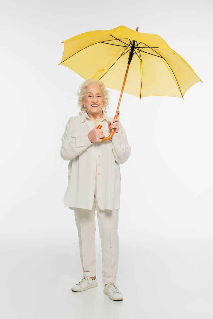 anciana sonriente con ropa casual de pie con paraguas amarillo sobre blanco - Foto, imagen