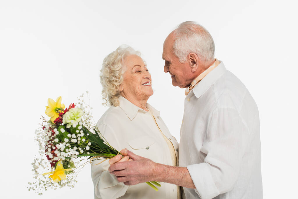Lächelndes älteres Paar, das einen Blumenstrauß in der Hand hält und einander isoliert auf weißem Grund ansieht - Foto, Bild