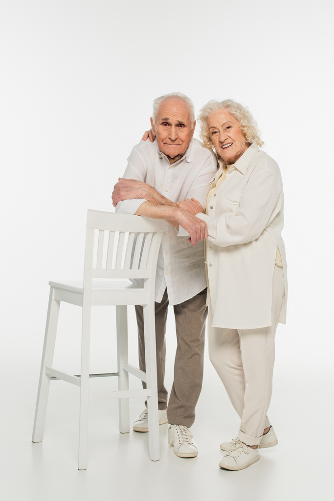 heureux couple âgé en vêtements décontractés debout près des selles et câlins sur blanc  - Photo, image