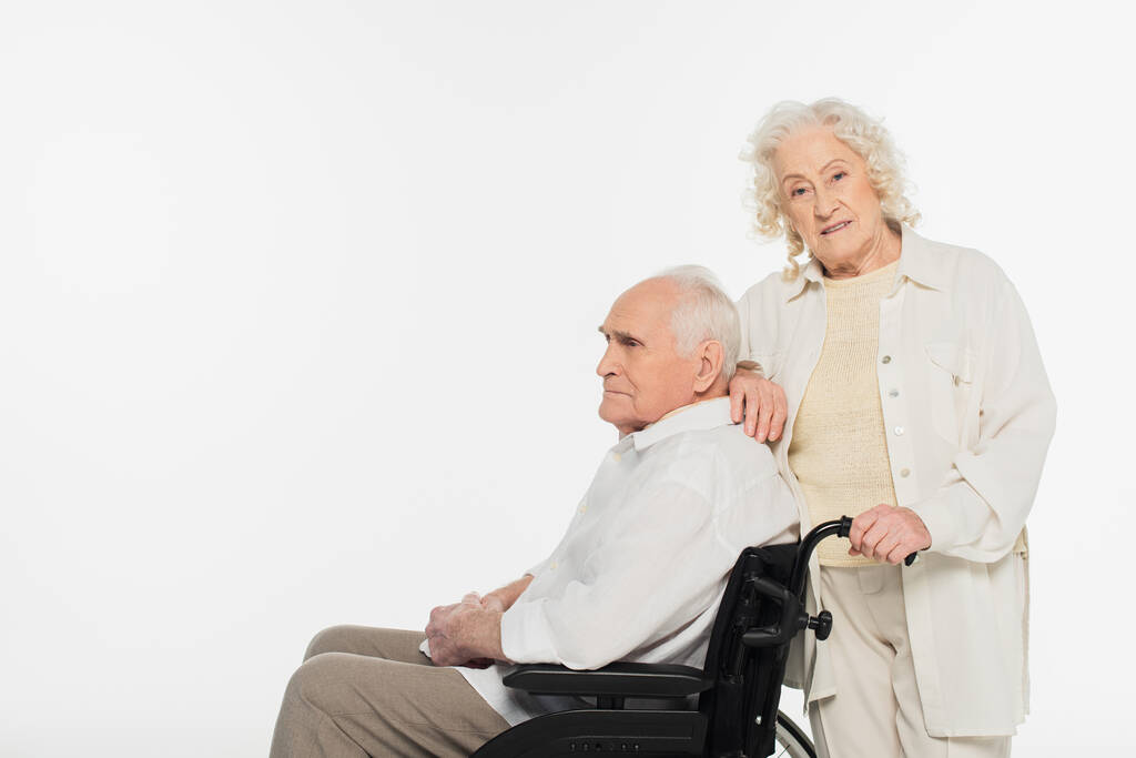 ηλικιωμένη γυναίκα στέκεται κοντά σύζυγος σε αναπηρικό καροτσάκι απομονωμένη σε λευκό - Φωτογραφία, εικόνα