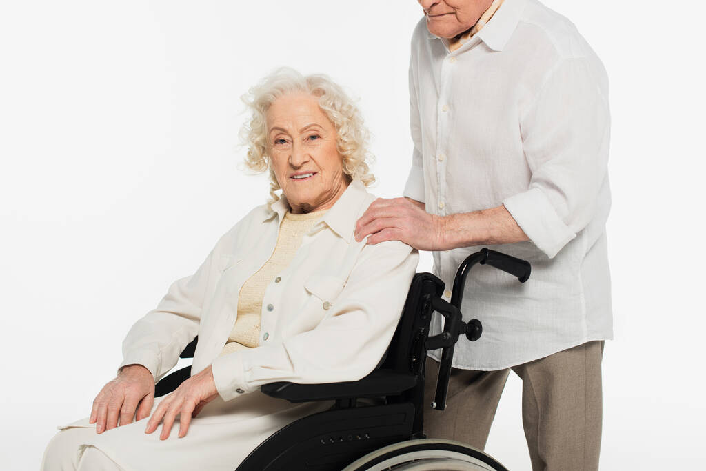 hymyilevä iäkäs nainen pyörätuolissa lähellä miestä pitämässä kädestä olkapäälle eristettynä valkoisella - Valokuva, kuva