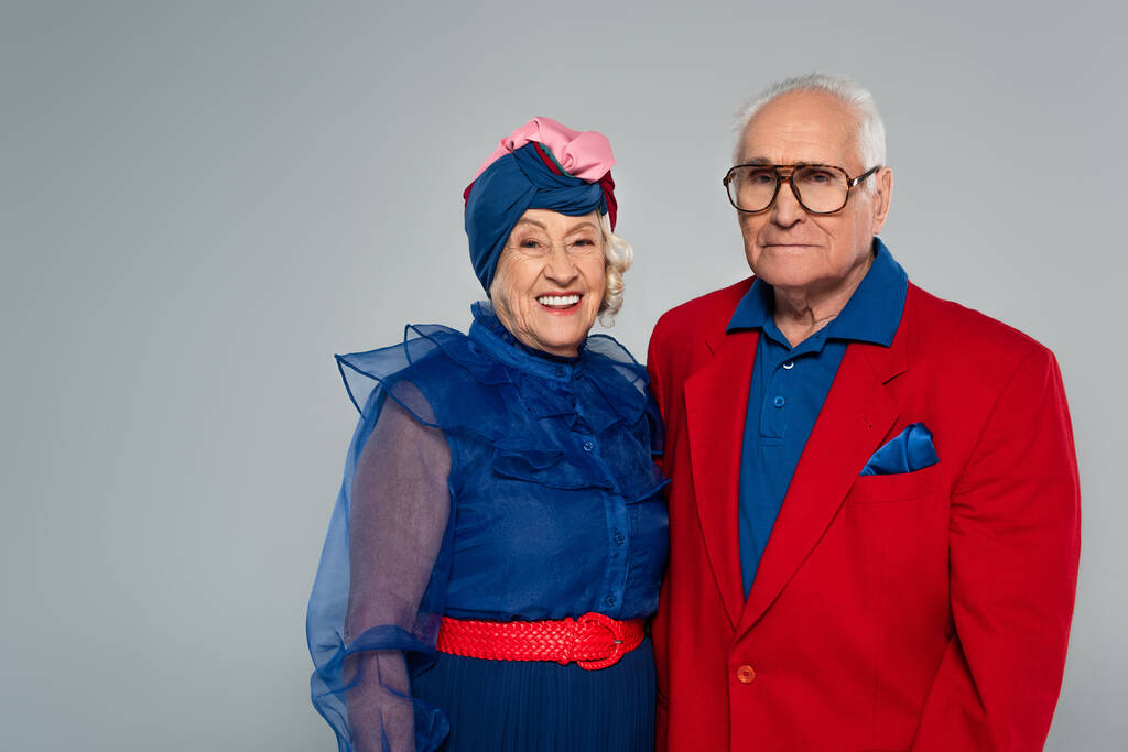 usmívající se starší pár ve stylovém oblečení při pohledu na kameru izolované na šedé - Fotografie, Obrázek