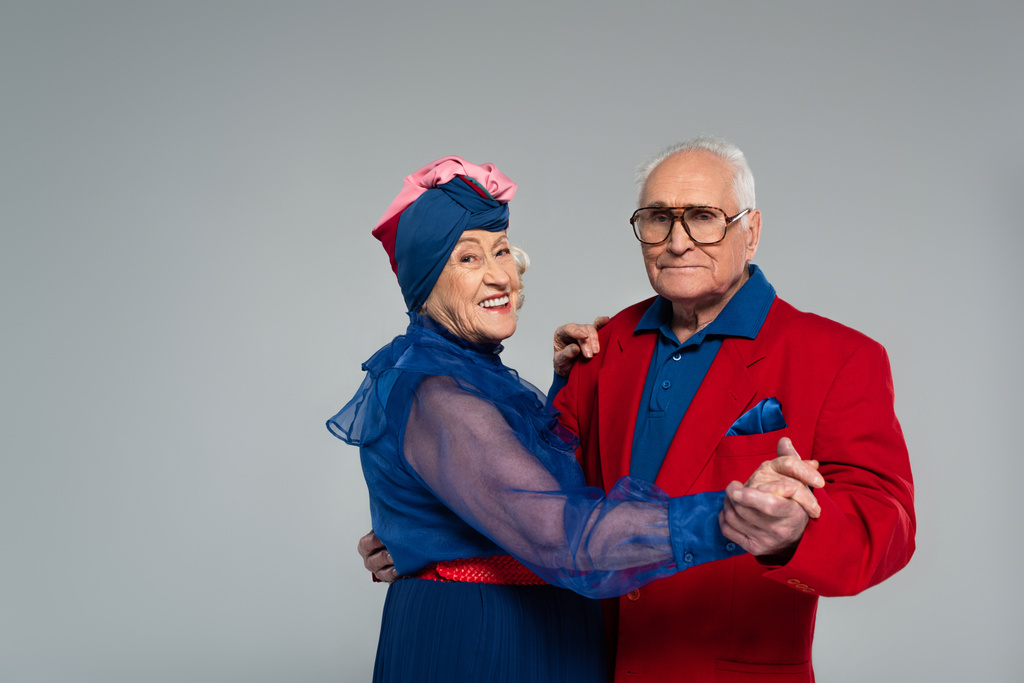 sonriente pareja de ancianos en vestido azul y chaqueta roja bailando aislados en gris - Foto, imagen