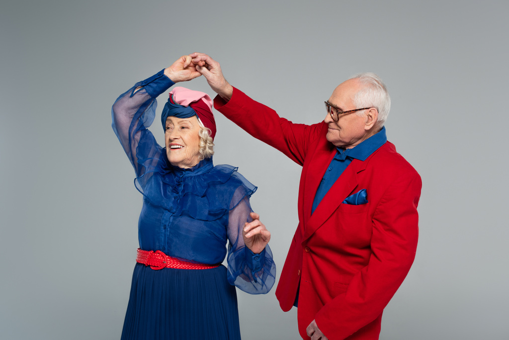 χαμογελαστό ηλικιωμένο ζευγάρι με μπλε φόρεμα και κόκκινο σακάκι που χορεύει απομονωμένο σε γκρι - Φωτογραφία, εικόνα