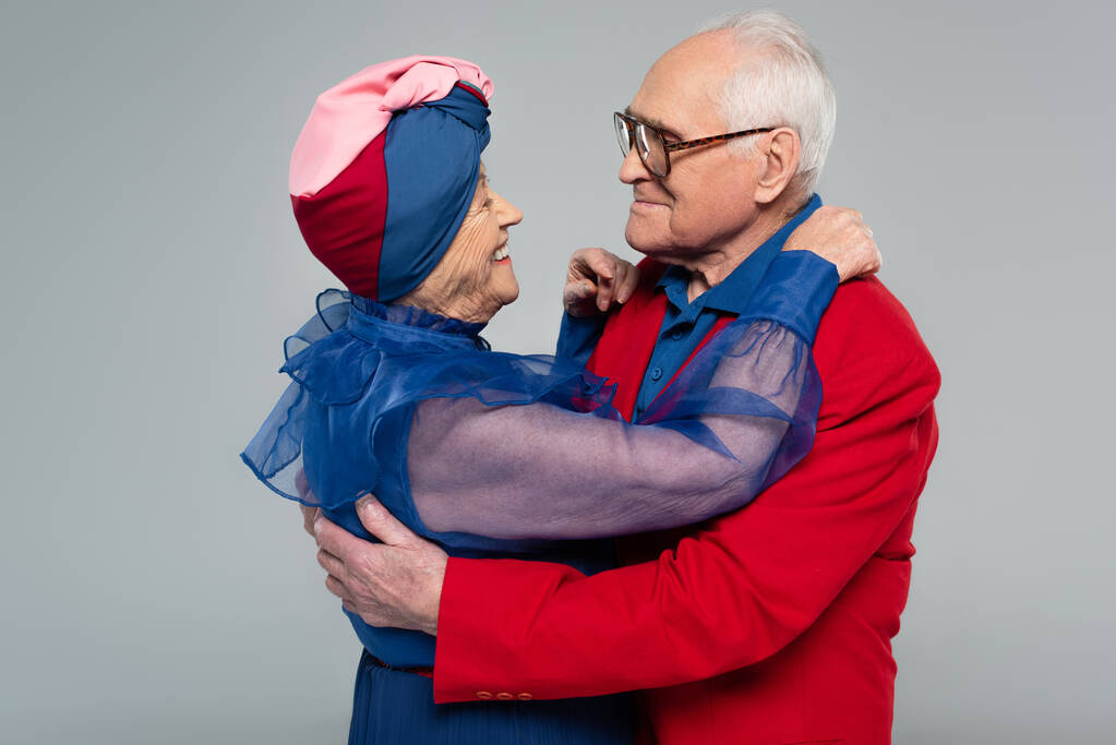 glimlachend ouder paar in blauwe jurk en rode blazer knuffelen geïsoleerd op grijs - Foto, afbeelding
