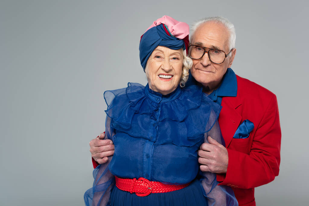 χαμογελαστός ηλικιωμένος άνδρας με κόκκινο σακάκι αγκαλιάζει τη σύζυγο με μπλε φόρεμα και τουρμπάνι απομονωμένο σε γκρι - Φωτογραφία, εικόνα