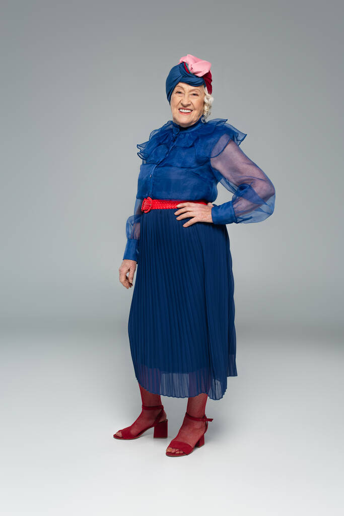 femme âgée souriante en robe bleue et turban posant avec la main sur la hanche sur gris - Photo, image