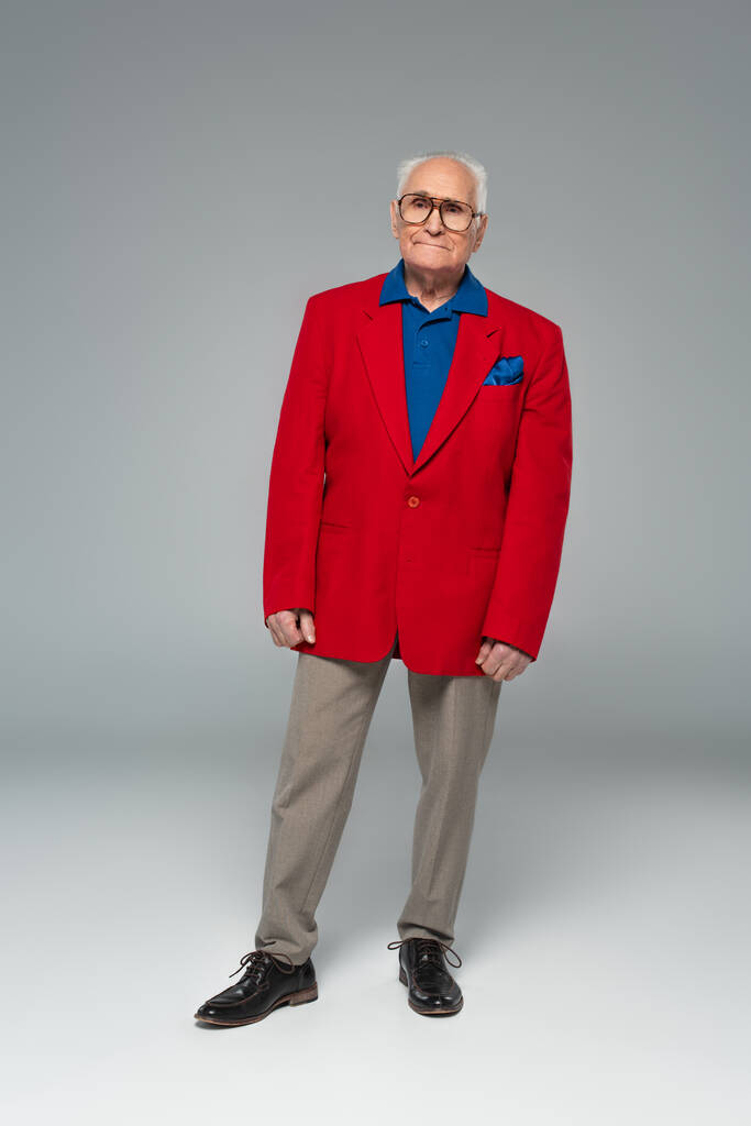 Вид в полный рост серьезного пожилого человека в очках, красном блейзере и синей рубашке, стоящего на сером - Фото, изображение