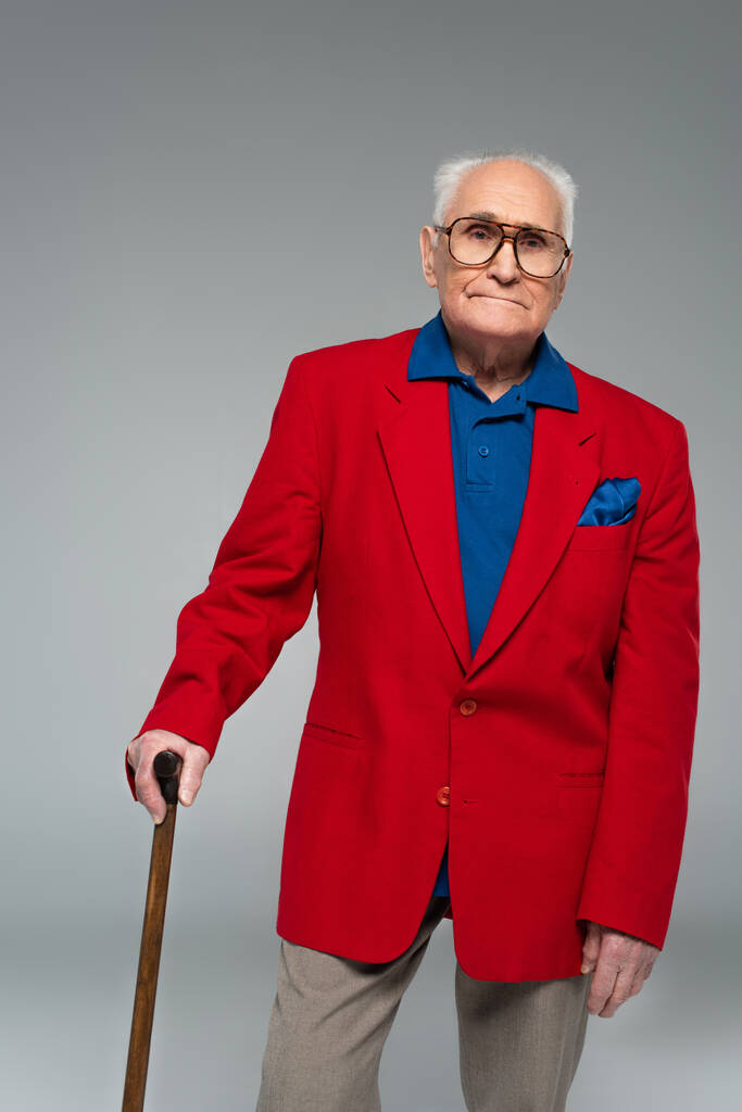 ernstige oudere man in rode blazer en bril staan met wandelstok geïsoleerd op grijs - Foto, afbeelding