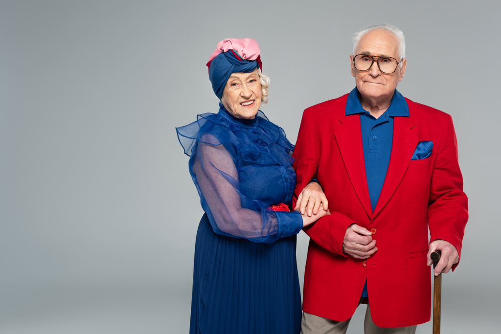 usmívající se starší žena v modrých šatech objímající manžel v červeném blejzru s vycházkovou holí izolované na šedé - Fotografie, Obrázek