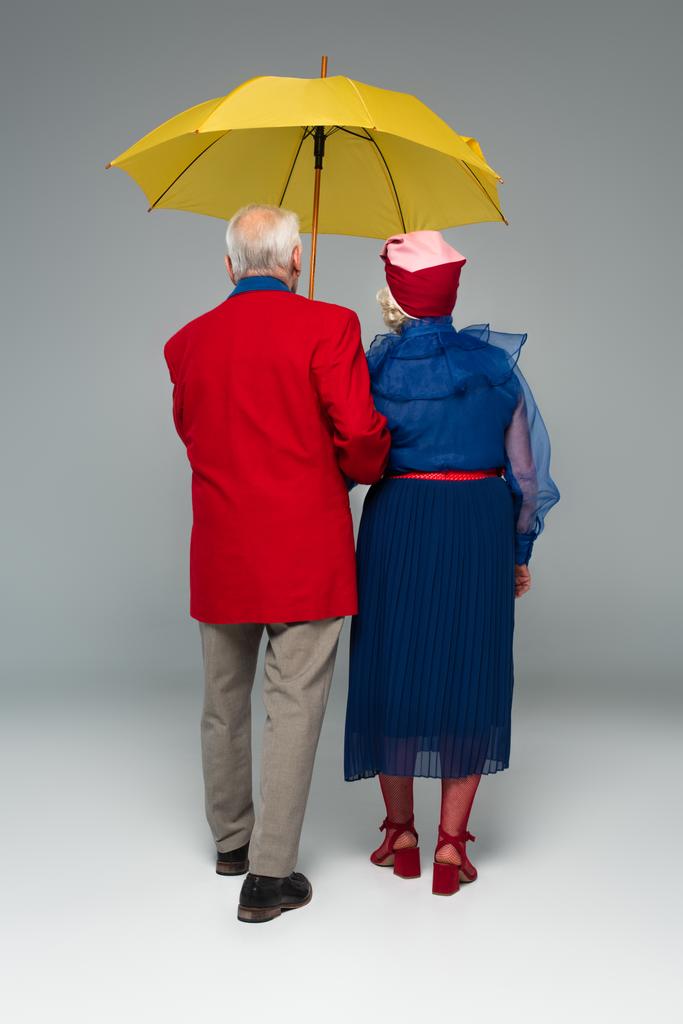 πίσω όψη του ηλικιωμένου ζευγαριού σε κομψά ρούχα περπάτημα με κίτρινη ομπρέλα σε γκρι - Φωτογραφία, εικόνα