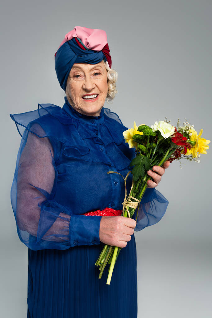 uśmiechnięta starsza kobieta w niebieskiej sukience i turban gospodarstwa bukiet kwiatów odizolowanych na szarości - Zdjęcie, obraz