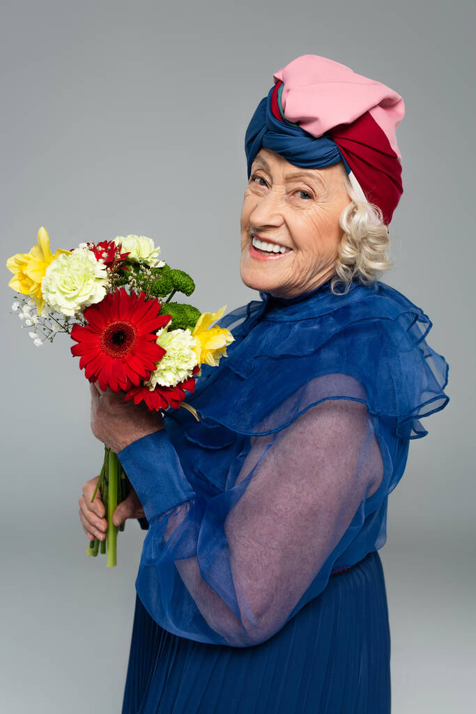 pozitív idős nő kék ruhában és turbán gazdaság csokor virág elszigetelt szürke - Fotó, kép