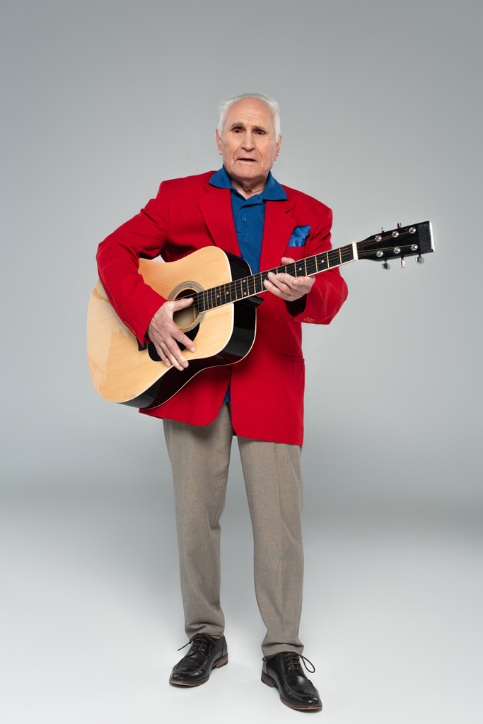 anciano en blazer rojo y pantalones marrones con guitarra acústica en gris - Foto, Imagen
