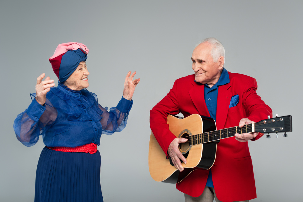 uśmiechnięty starzec w czerwonej marynarce gra na gitarze akustycznej w pobliżu tańczącej kobiety w niebieskiej sukience odizolowanej na szarości - Zdjęcie, obraz