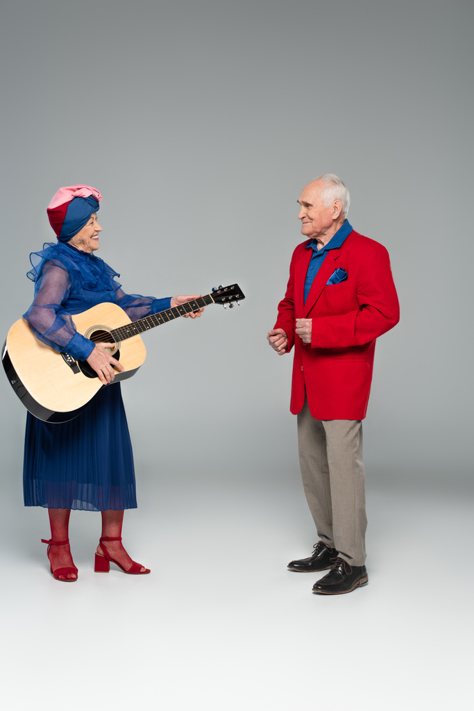 Positives älteres Paar in stylischer Kleidung tanzt mit Akustikgitarre auf grau - Foto, Bild