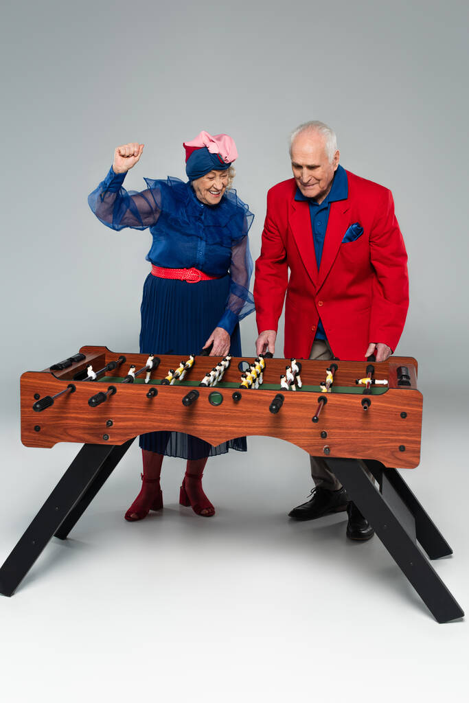 ενθουσιασμένοι ηλικιωμένοι ζευγάρι σε κομψά ρούχα παίζοντας ποδόσφαιρο τραπέζι με χειρονομία νίκη σε γκρι - Φωτογραφία, εικόνα