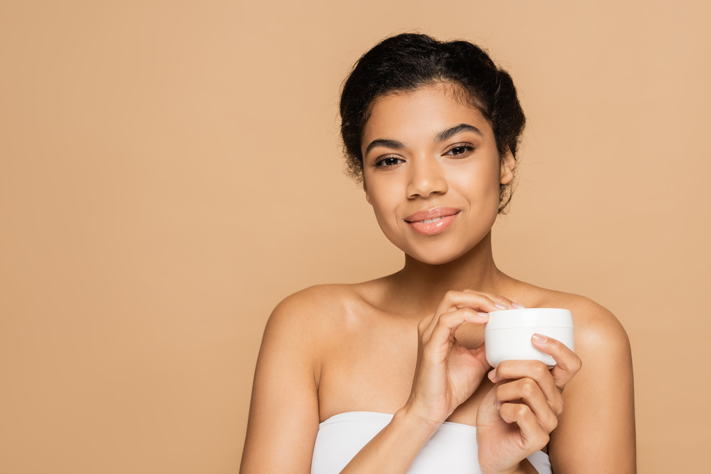 smiling african american woman with bare shoulders holding container with cosmetic cream isolated on beige  - Photo, Image