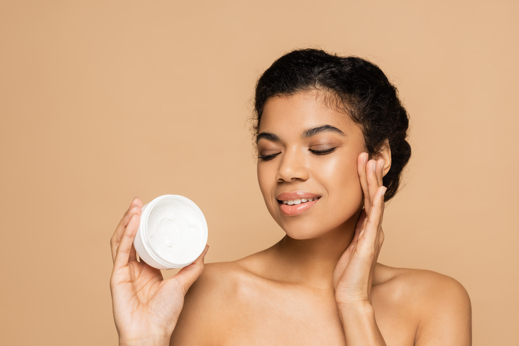 lächelnde Afroamerikanerin mit nackten Schultern beim Betrachten eines Behälters mit Kosmetikcreme isoliert auf beige  - Foto, Bild