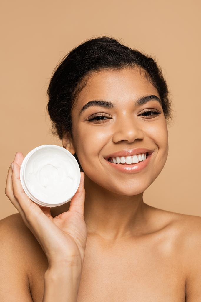 pleased african american woman with bare shoulders holding container with face cream isolated on beige  - Photo, Image
