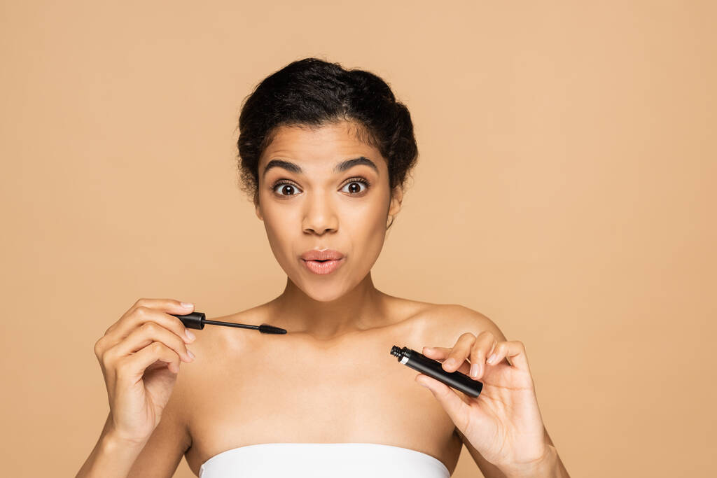 amazed african american woman with bare shoulders holding mascara tube and brush isolated on beige  - Photo, Image